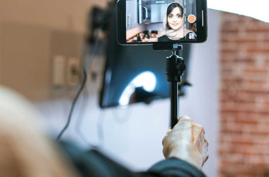 A smartphone mounted on a stick displays a woman smiling and ready to record a video
